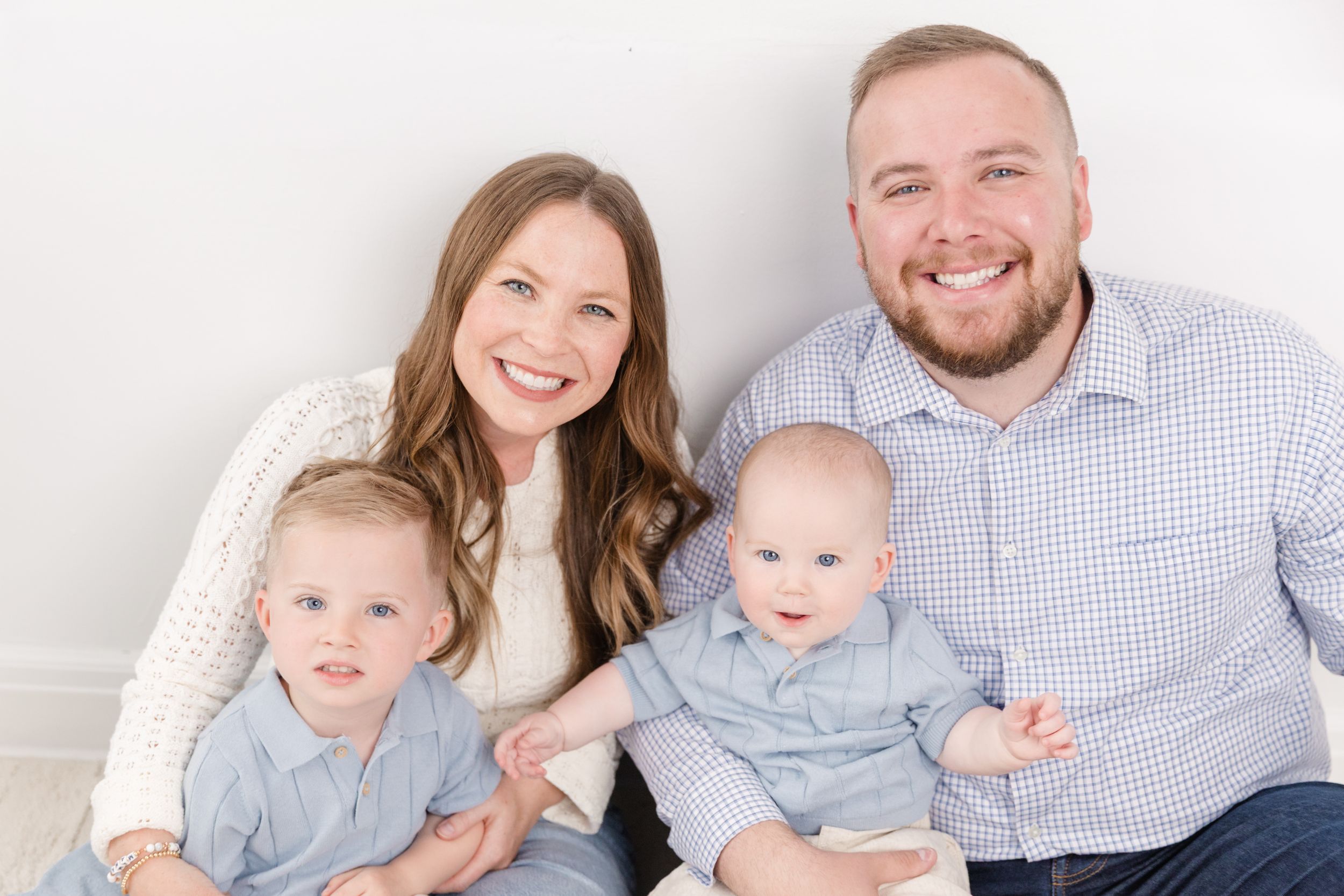 Happy family portrait with smiling parents and two young boys in light blue outfits against a white background.