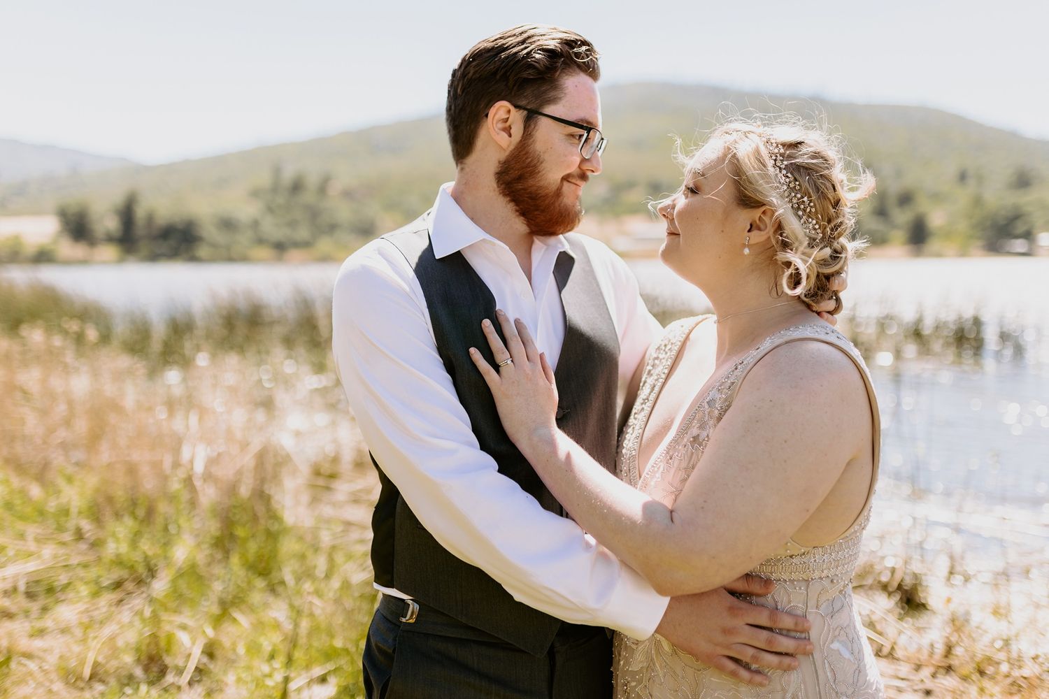 A couple in wedding attire embraces near a scenic lake with mountains in the background on a sunny day at Lake Cuyamaca, Julian.