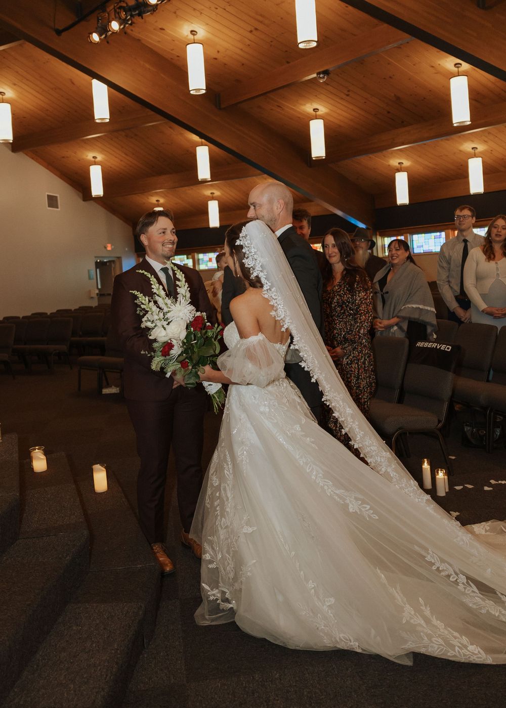 Bride walking down church aisle in lace gown with long veil toward smiling groom.