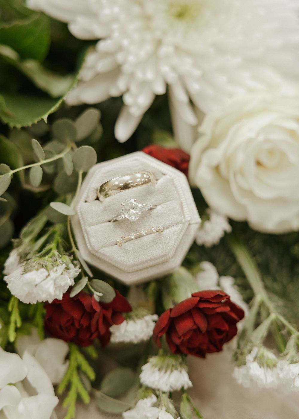 Diamond engagement ring and wedding bands displayed in velvet octagon box surrounded by red and white florals.