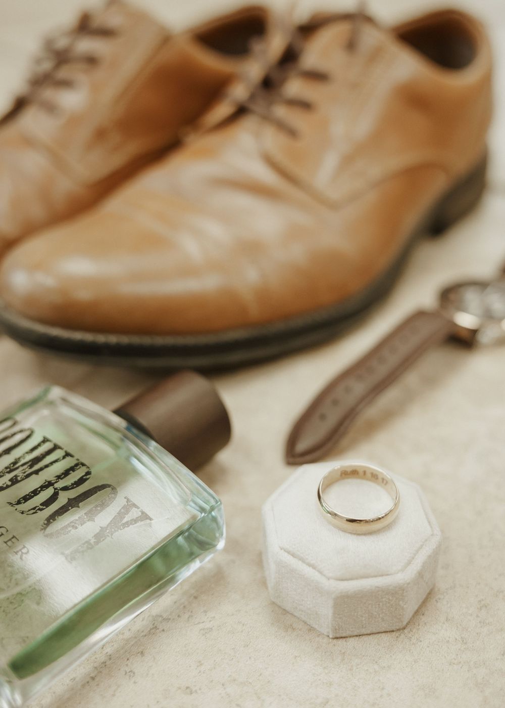 Groom wedding details flat lay with tan oxford shoes, cologne, watch strap, and gold band in ring box.