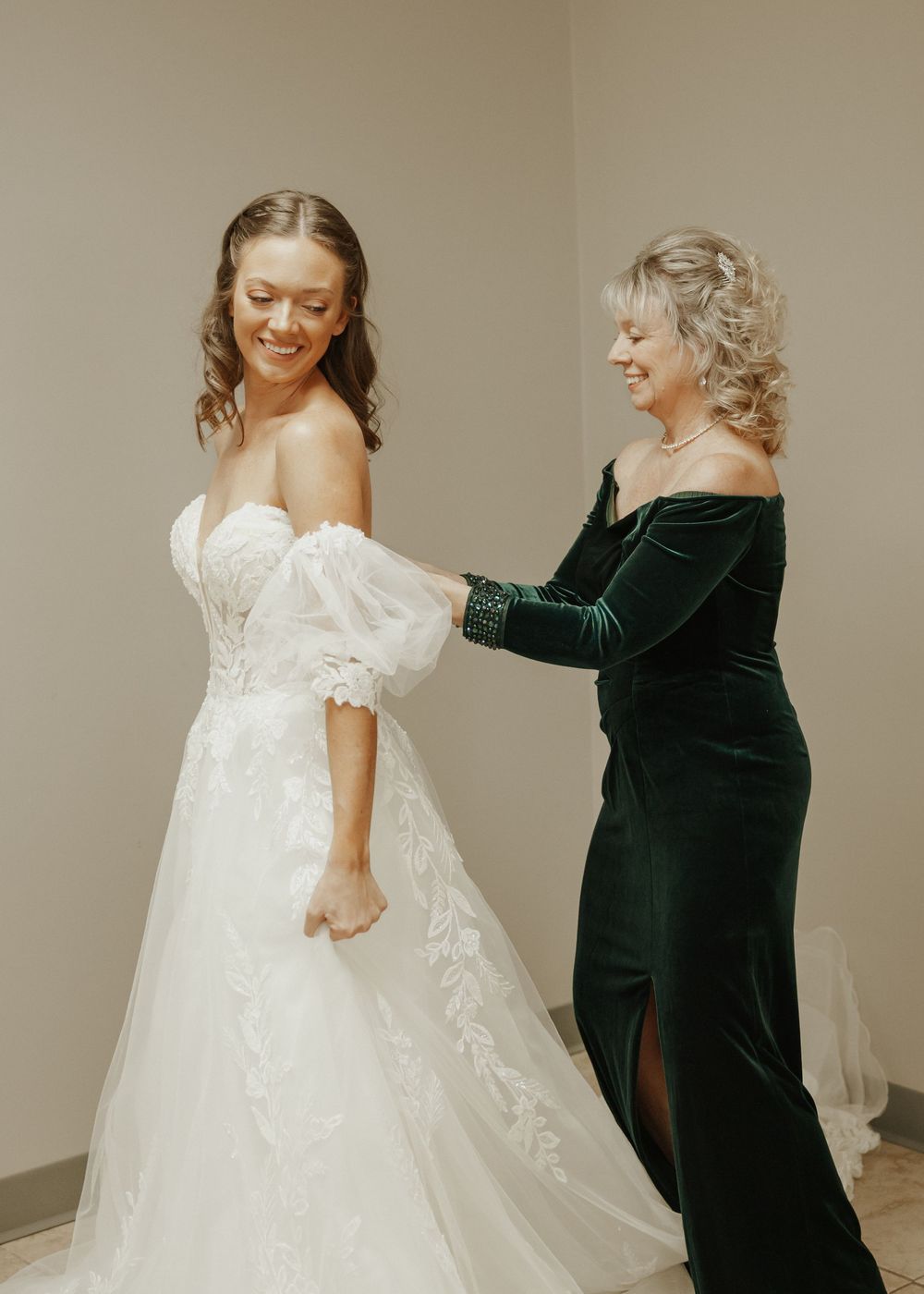 Smiling bride in lace wedding gown being helped by woman in green velvet dress.