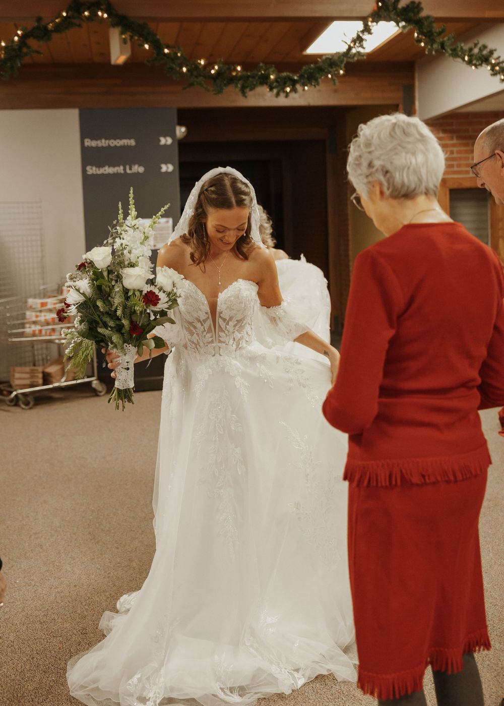 Bride in lace wedding gown with veil holding white bouquet greeted by elderly woman.