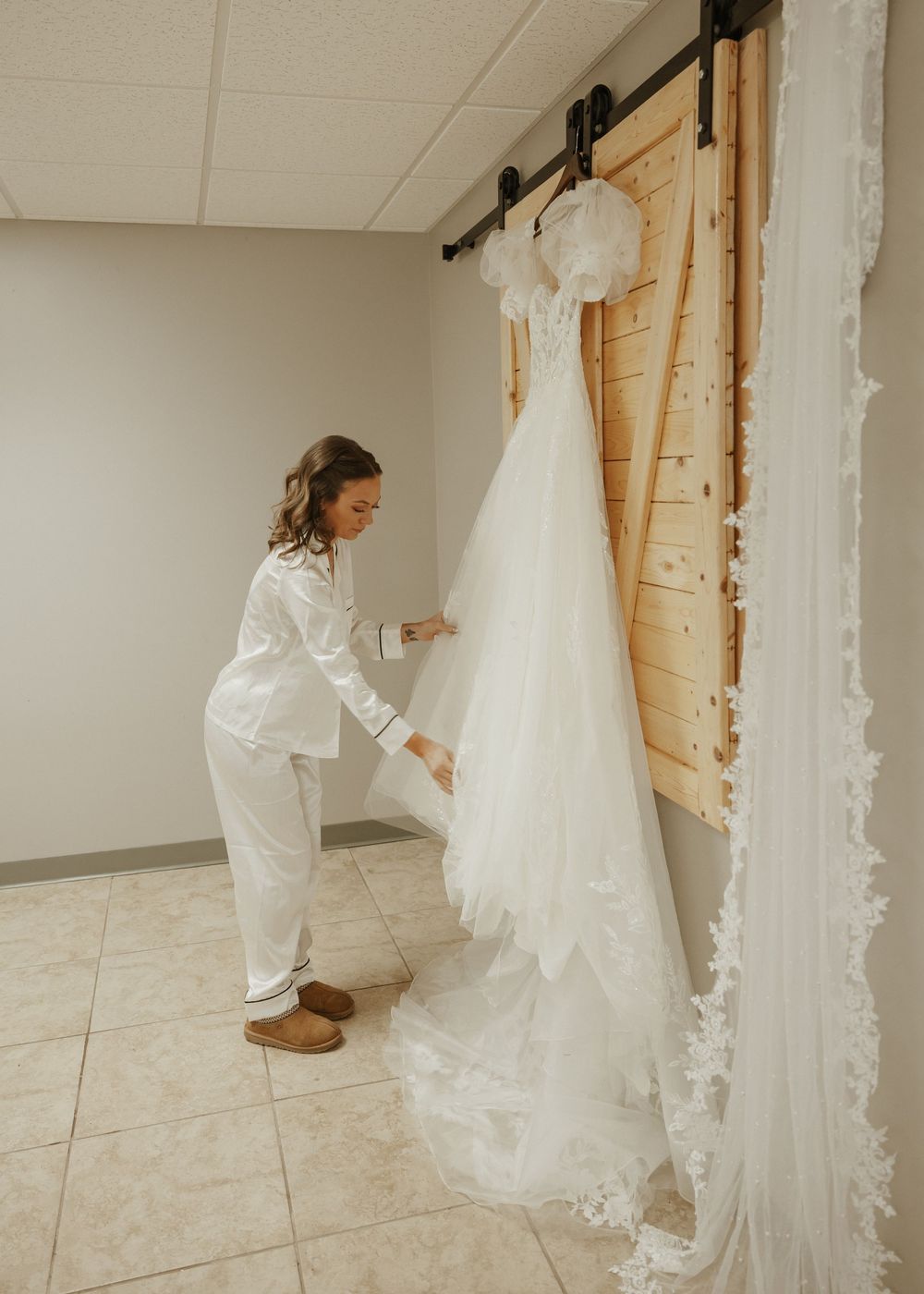 Bride in white pajamas examining lace wedding gown hanging on rustic barn door.
