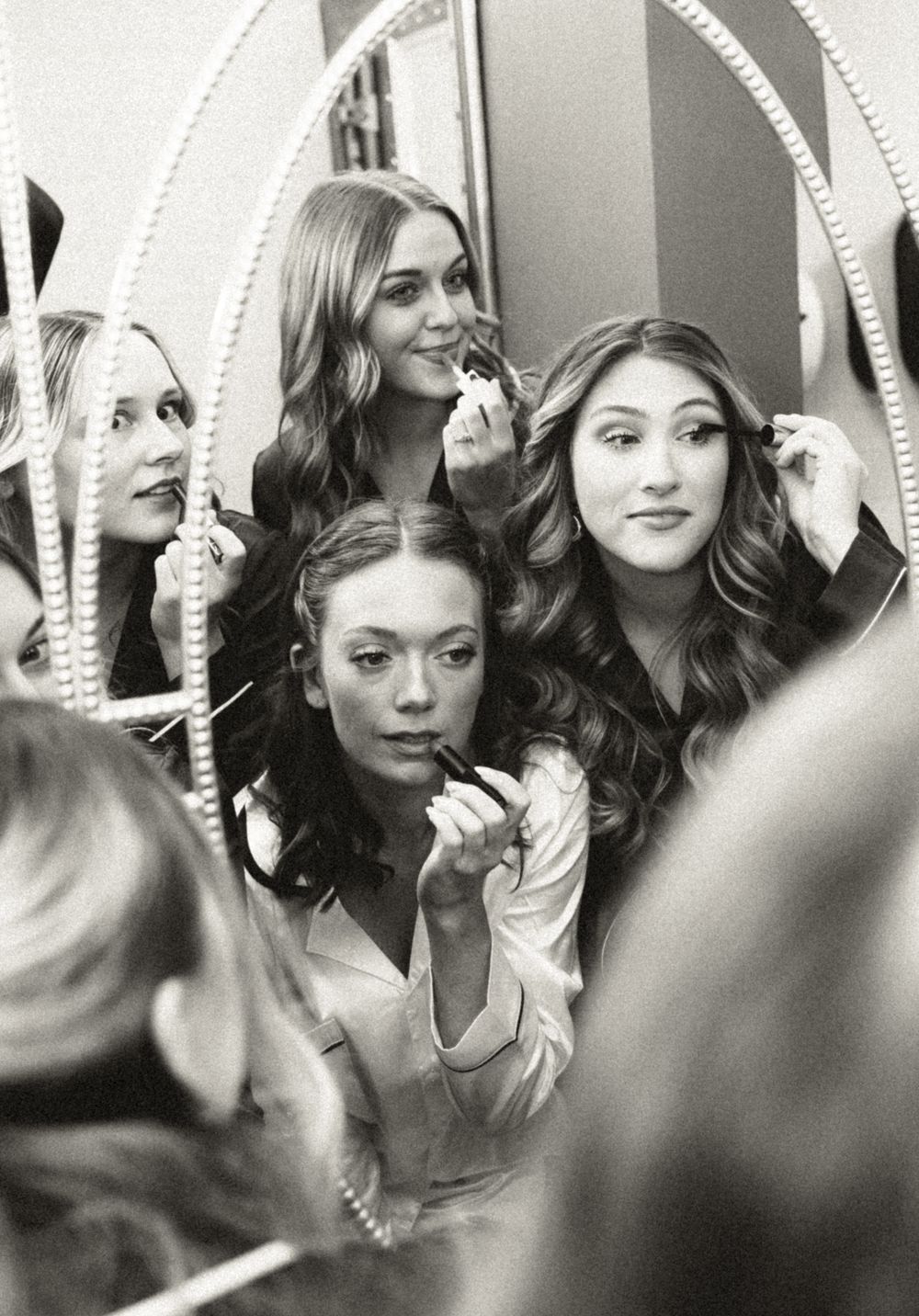 Bridesmaids applying makeup together in mirror reflection in black and white photo.