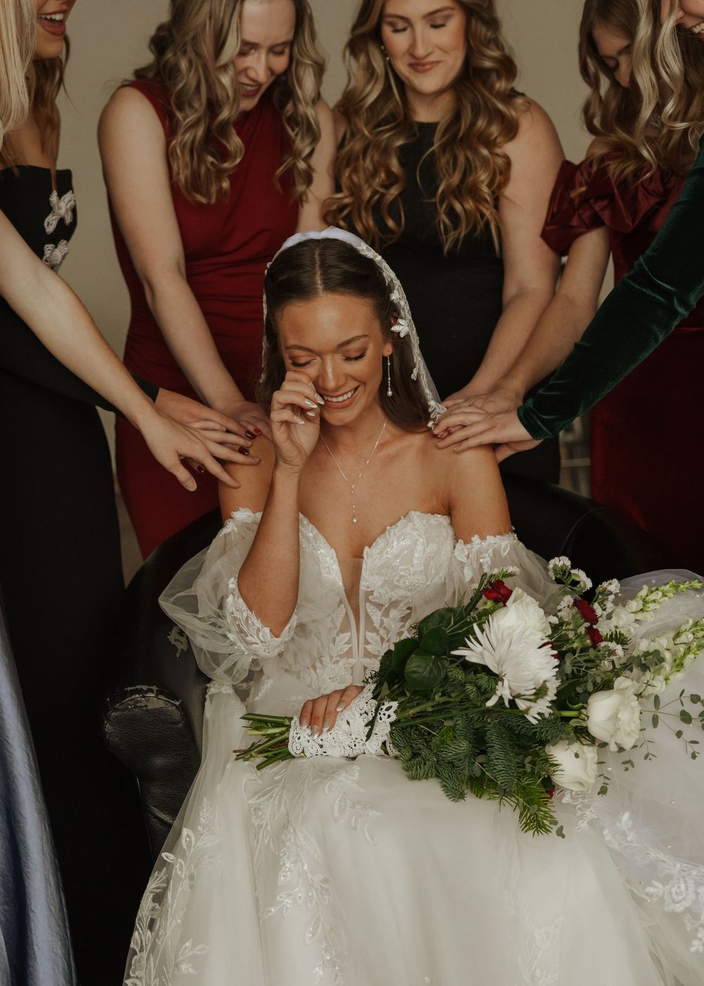 Emotional bride in lace gown wiping tears while bridesmaids in red and green surround her.