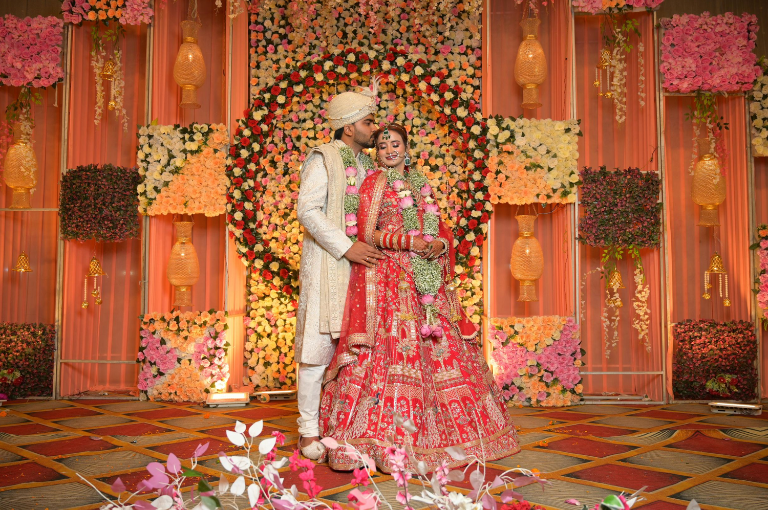 Elegant Indian wedding mandap decorated with pink and orange flowers, crystal chandeliers, and warm lighting.