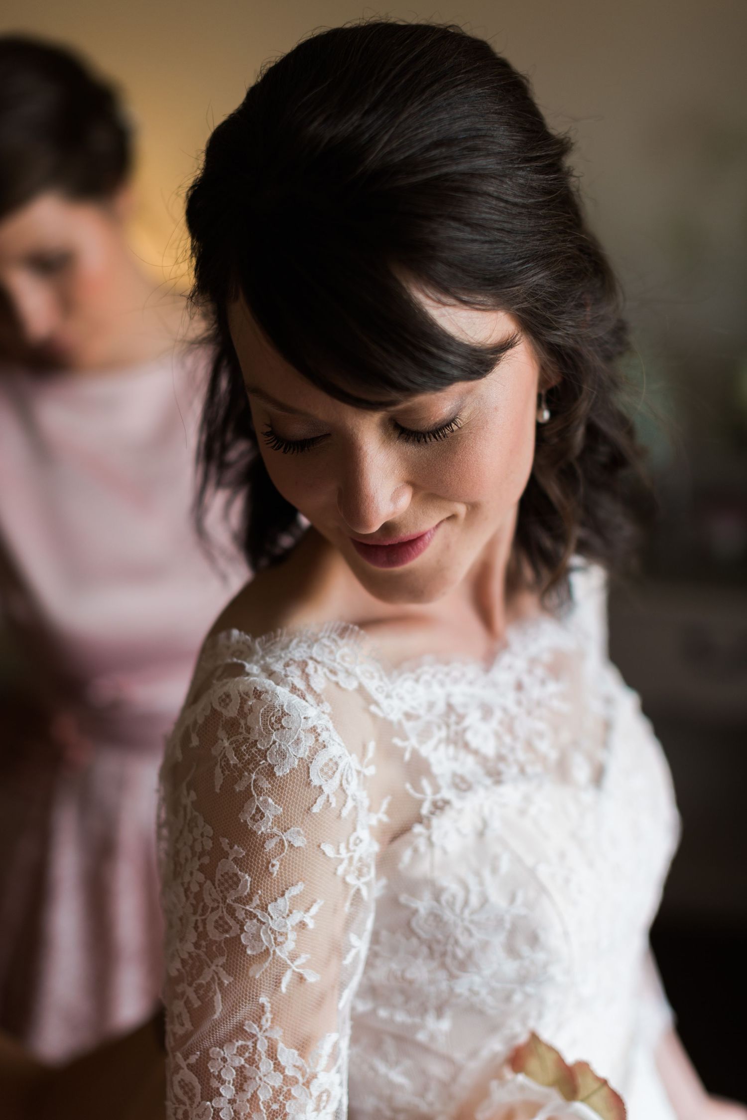 Bride in lace long-sleeve wedding gown looking down while bridesmaid assists with dress in soft natural light.