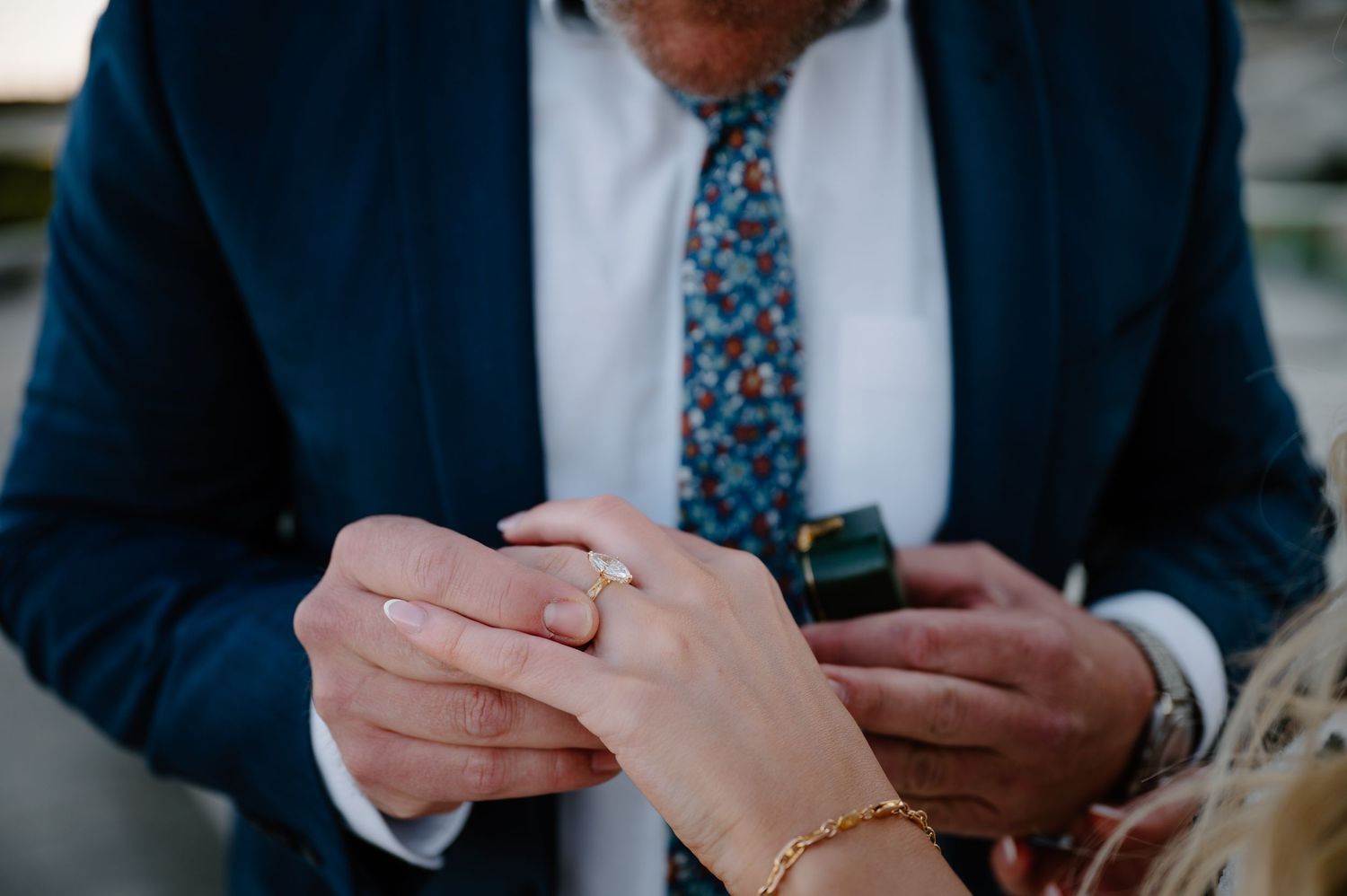 Man proposes on one knee to woman in white dress on steps of historic building with clock tower.