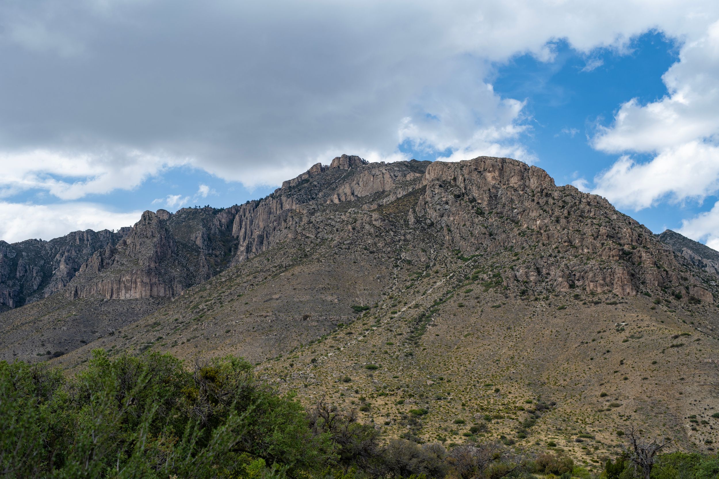 Rocky mountain peak with rugged cliffs, desert shrubs, and green trees under a partly cloudy blue sky.