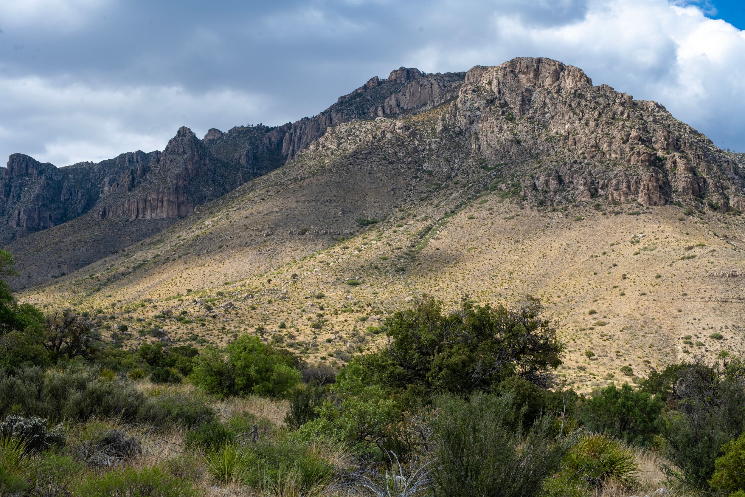 Rocky mountain landscape with desert scrub vegetation under cloudy skies in an arid southwestern wilderness.