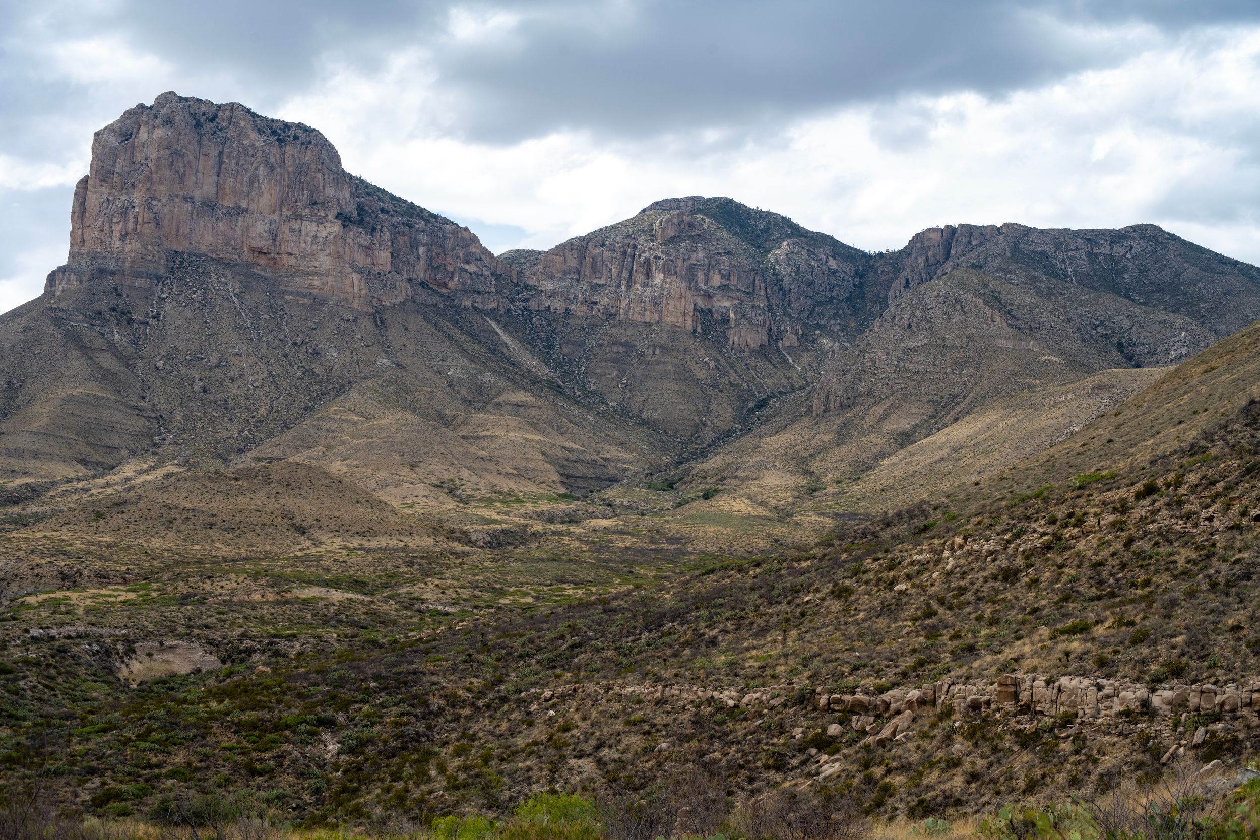 Rugged desert mountains with rocky cliffs rise above a scrubby valley under cloudy skies in a dramatic arid landscape.