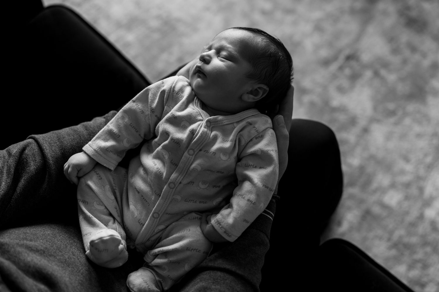 A sleeping baby in light colored sleepwear rests peacefully on a dark couch in black and white photography.