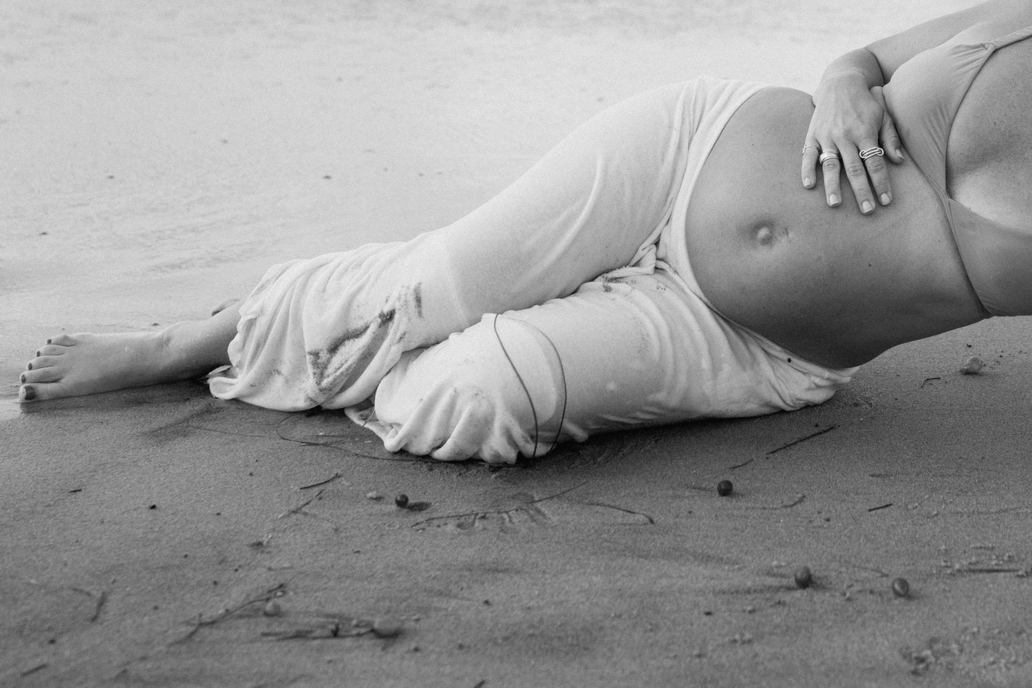Black and white photo of a pregnant woman lying on a sandy beach, cradling her bare baby bump.