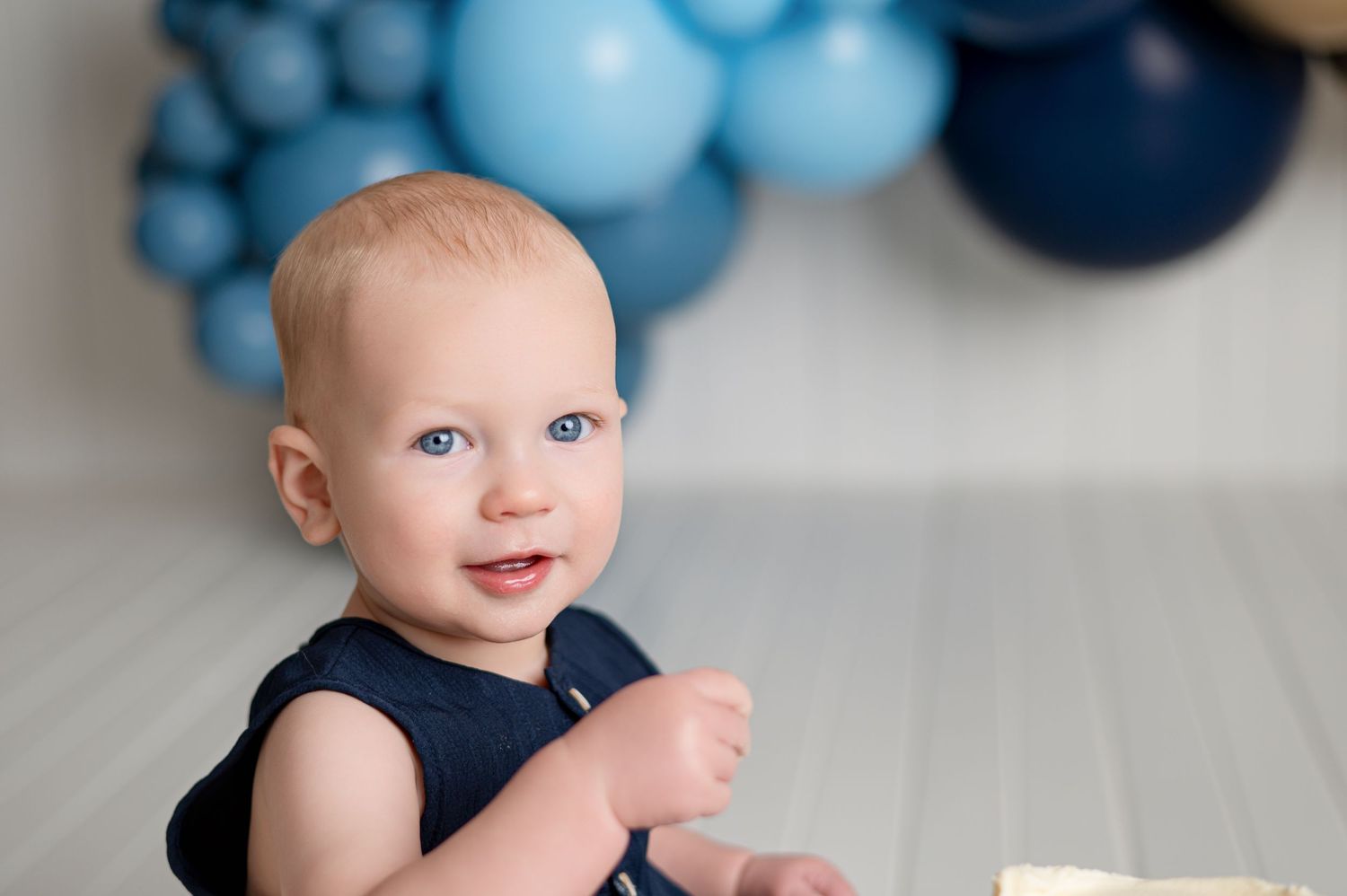Smiling toddler in teal romper sits on white floor with blue, white, and silver balloon garland backdrop.