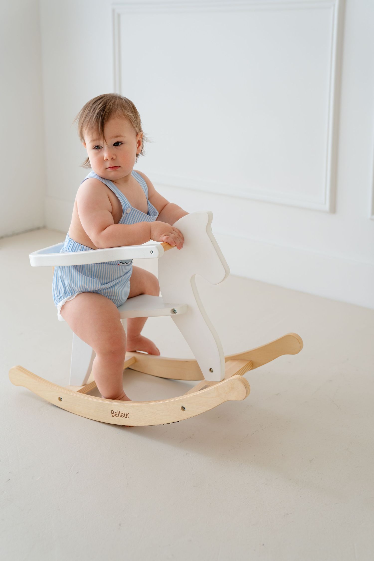 A baby in a blue striped romper stands on a white wooden rocking horse toy in a bright white room.