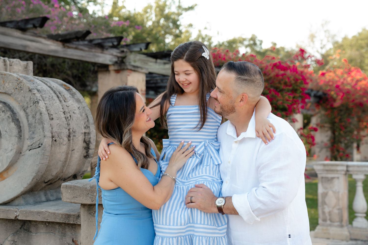 Happy family of three sharing a joyful moment outdoors, with blooming flowers and stone columns in the background.