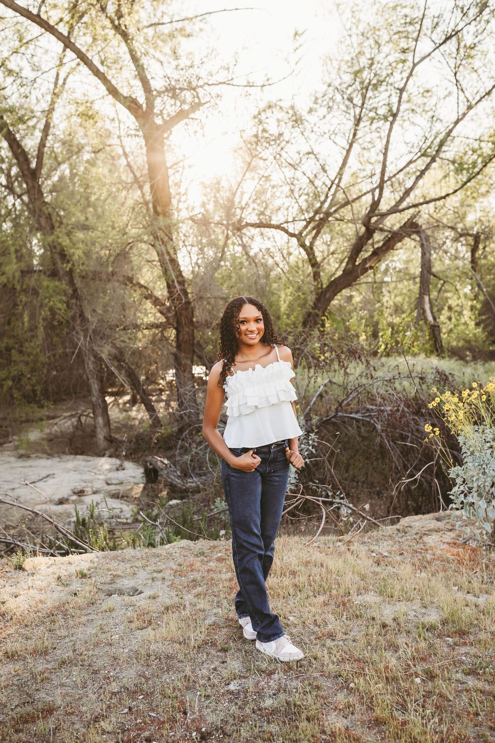 Young woman in white ruffled top and jeans smiling outdoors in a sunlit wooded setting.