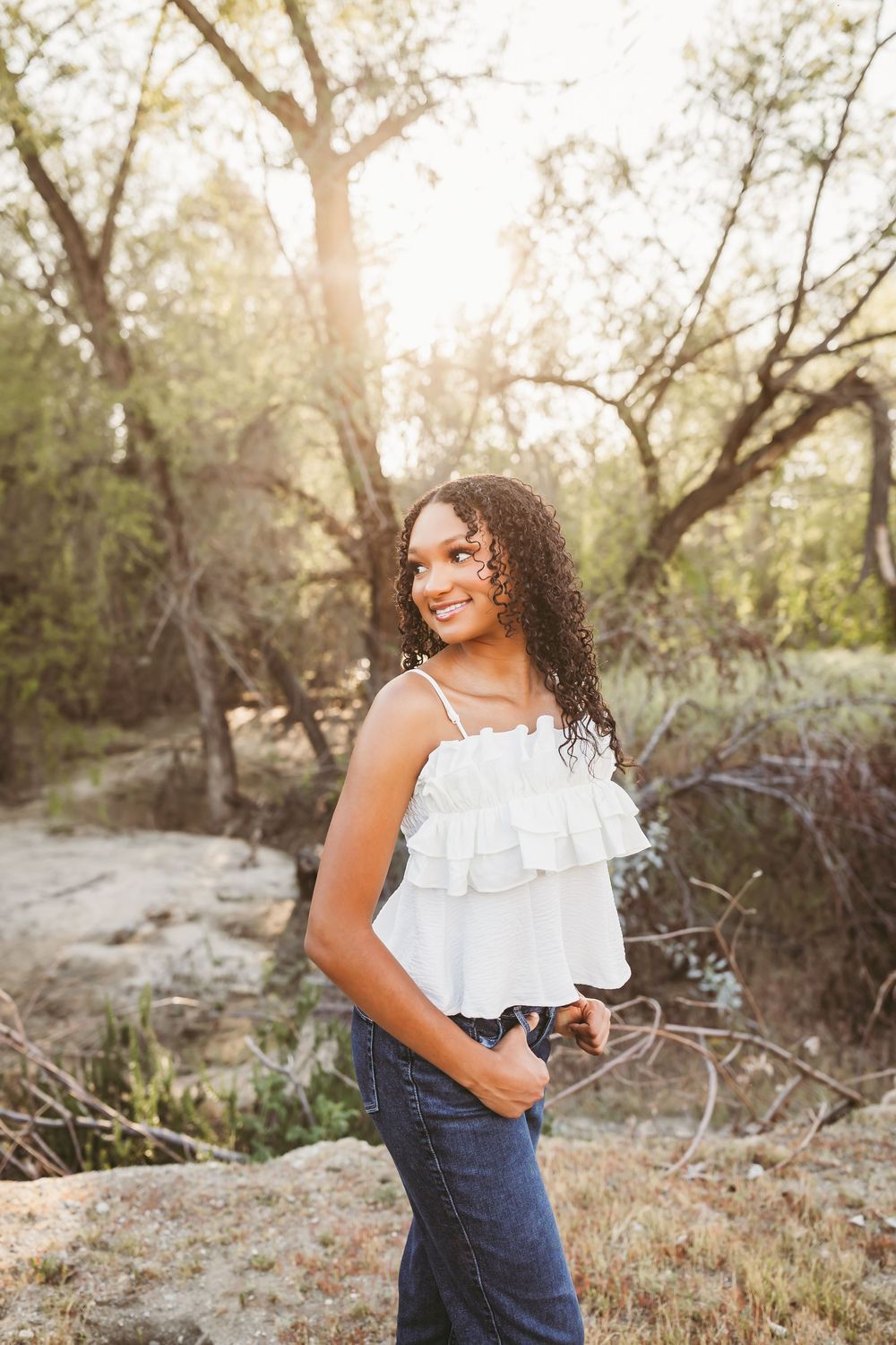 Young woman with curly hair smiles in white ruffled top and jeans, standing outdoors among trees in golden sunlight.