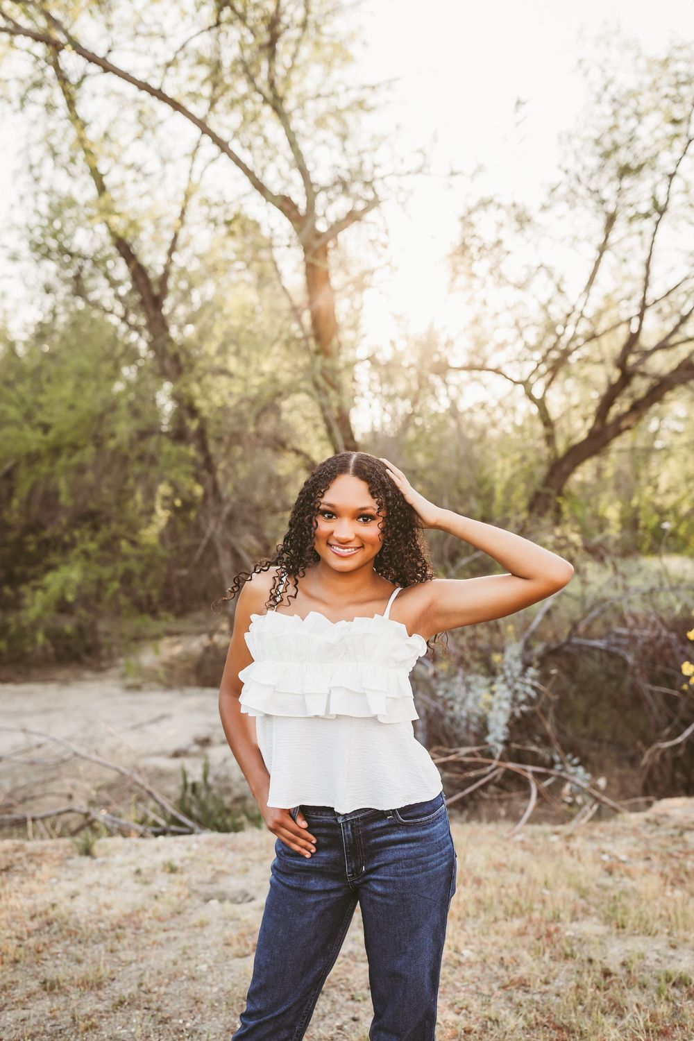 Young woman in white ruffled top and jeans smiling outdoors in golden sunlight among trees.