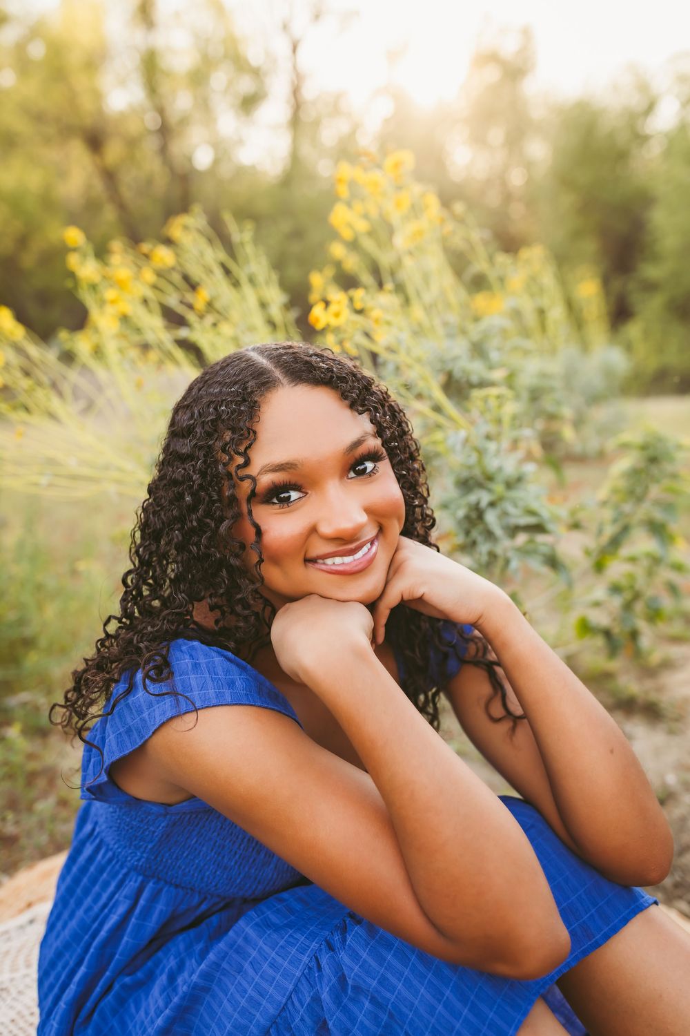 Young woman in blue dress smiling outdoors with yellow wildflowers in sunlit background.