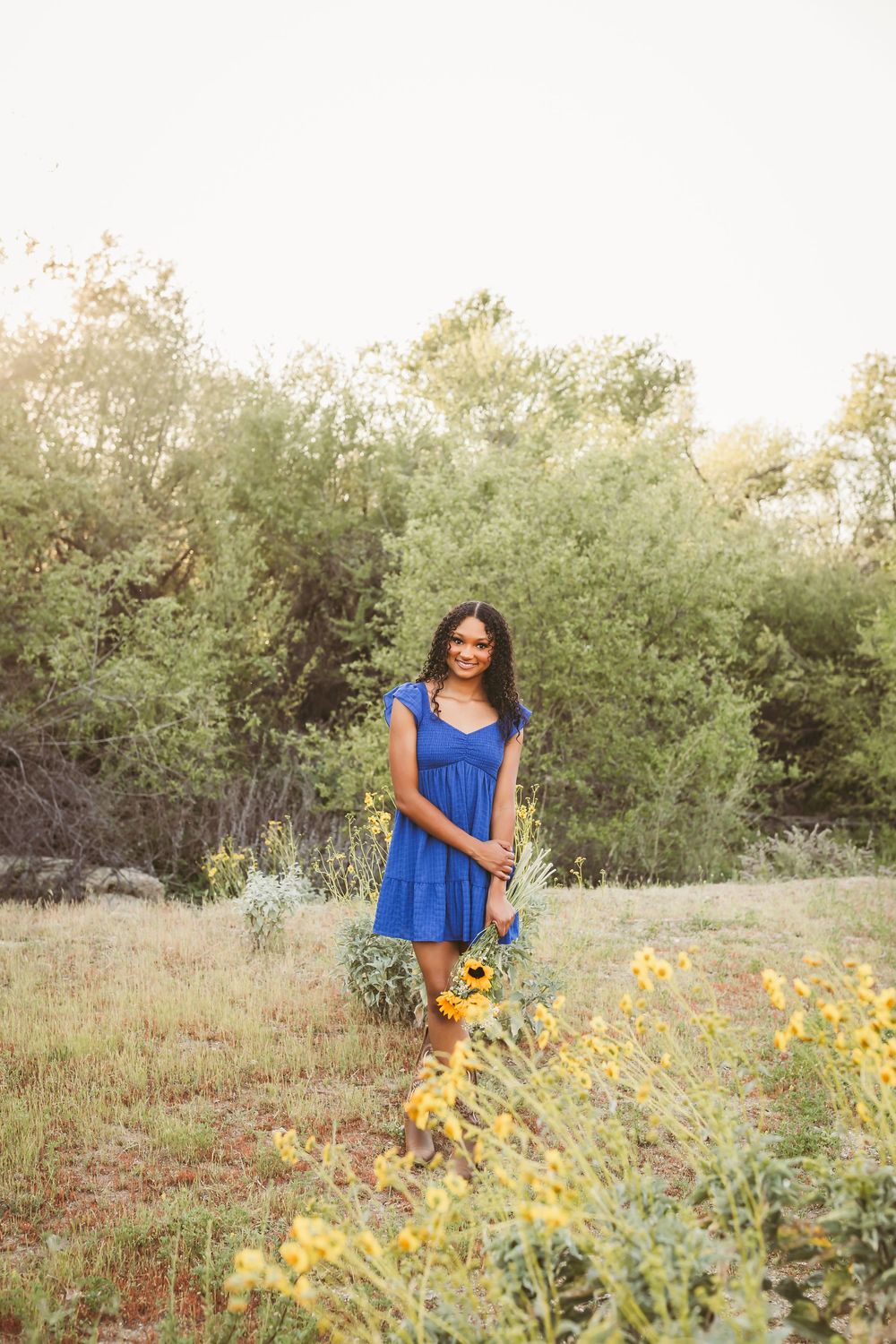 Young woman in blue dress holding sunflowers in a wildflower field with green trees backdrop.