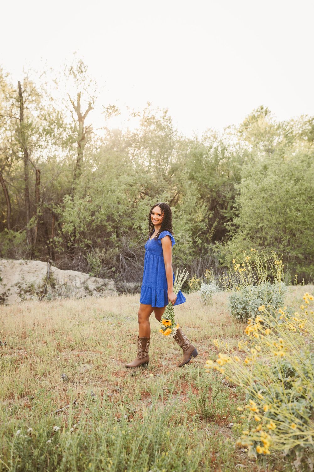 Woman in blue dress and cowboy boots holding wildflowers in a sunlit meadow surrounded by green trees.