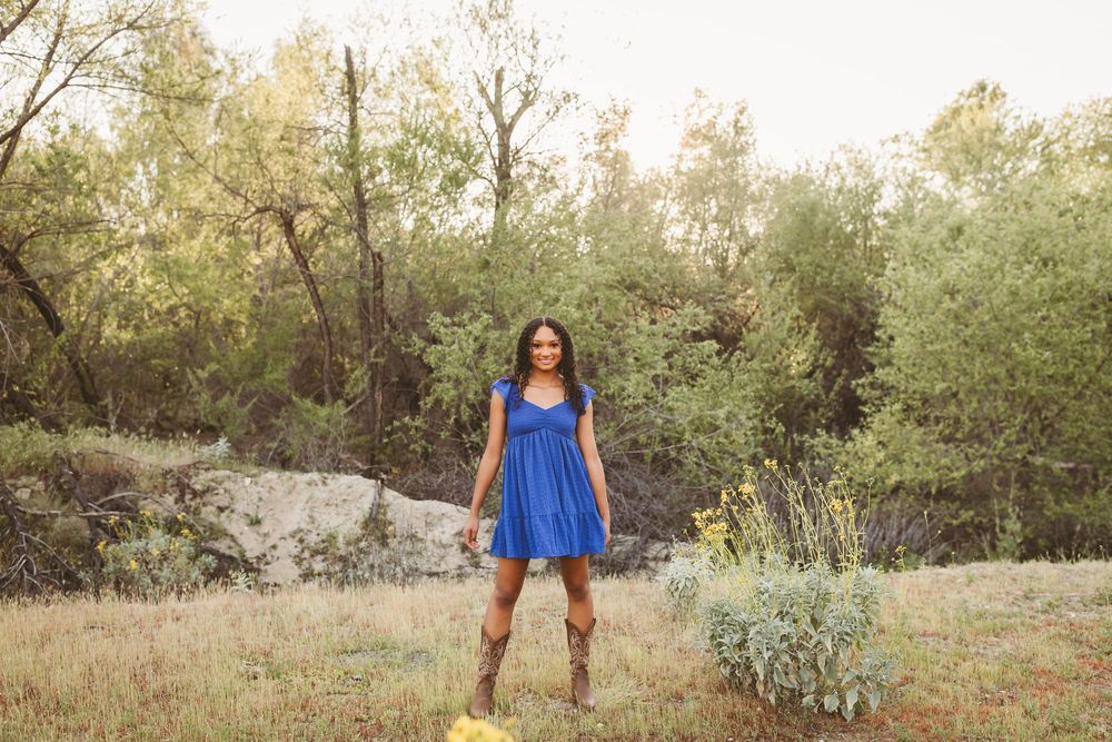 A young woman in a blue dress and cowboy boots stands smiling in a natural outdoor setting with trees and rocks.