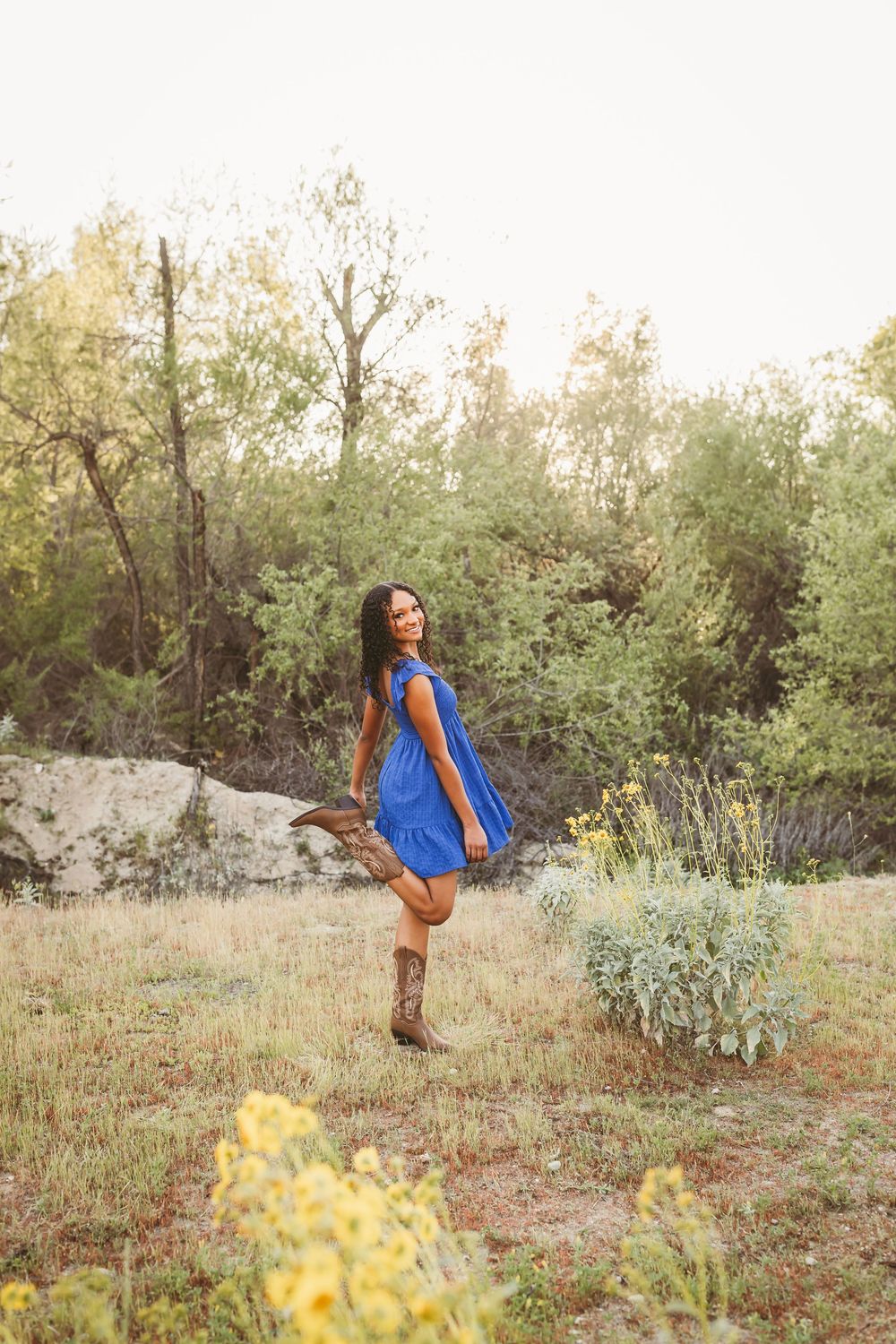 Young woman in blue dress and cowboy boots posing playfully in a sunlit field with wildflowers.