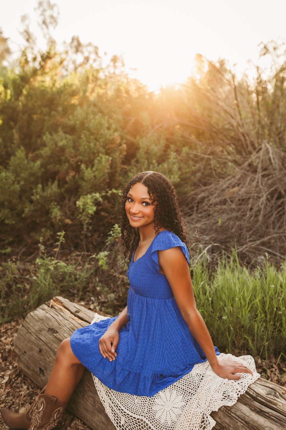 Young woman in blue dress sitting on a log with golden sunset light in a lush outdoor setting.