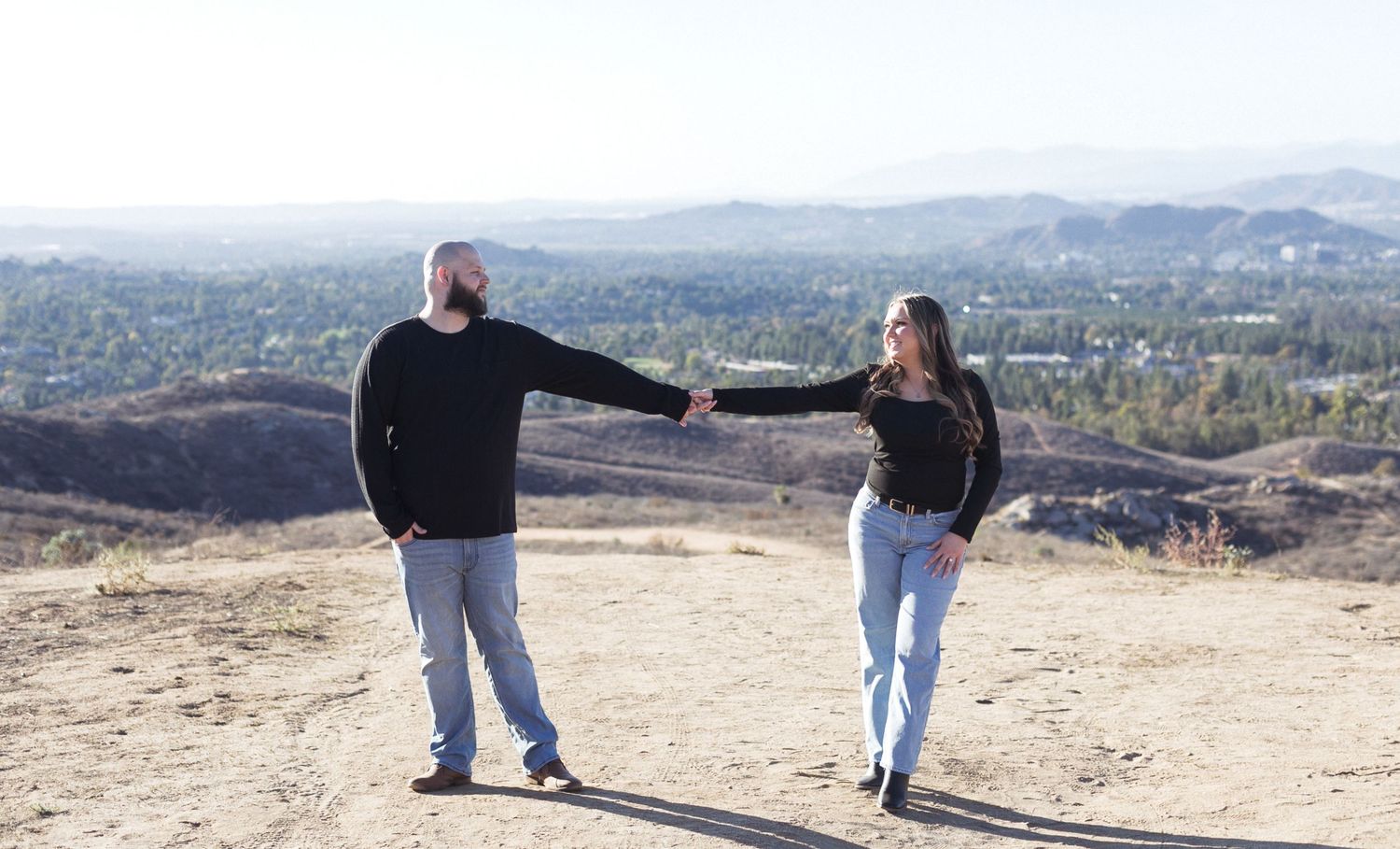 Couple holding hands on a hilltop with a scenic valley and mountain view in the background on a sunny day.