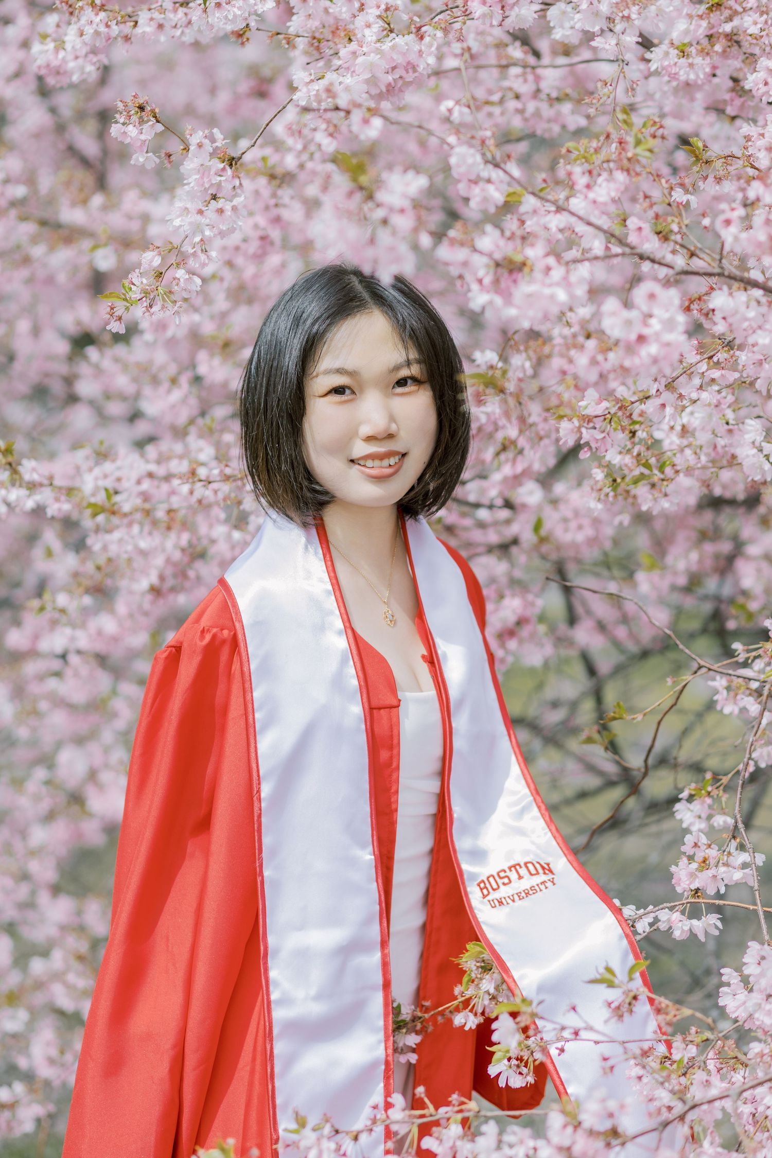 Boston University graduate smiles in red cap and gown among pink cherry blossoms in spring.