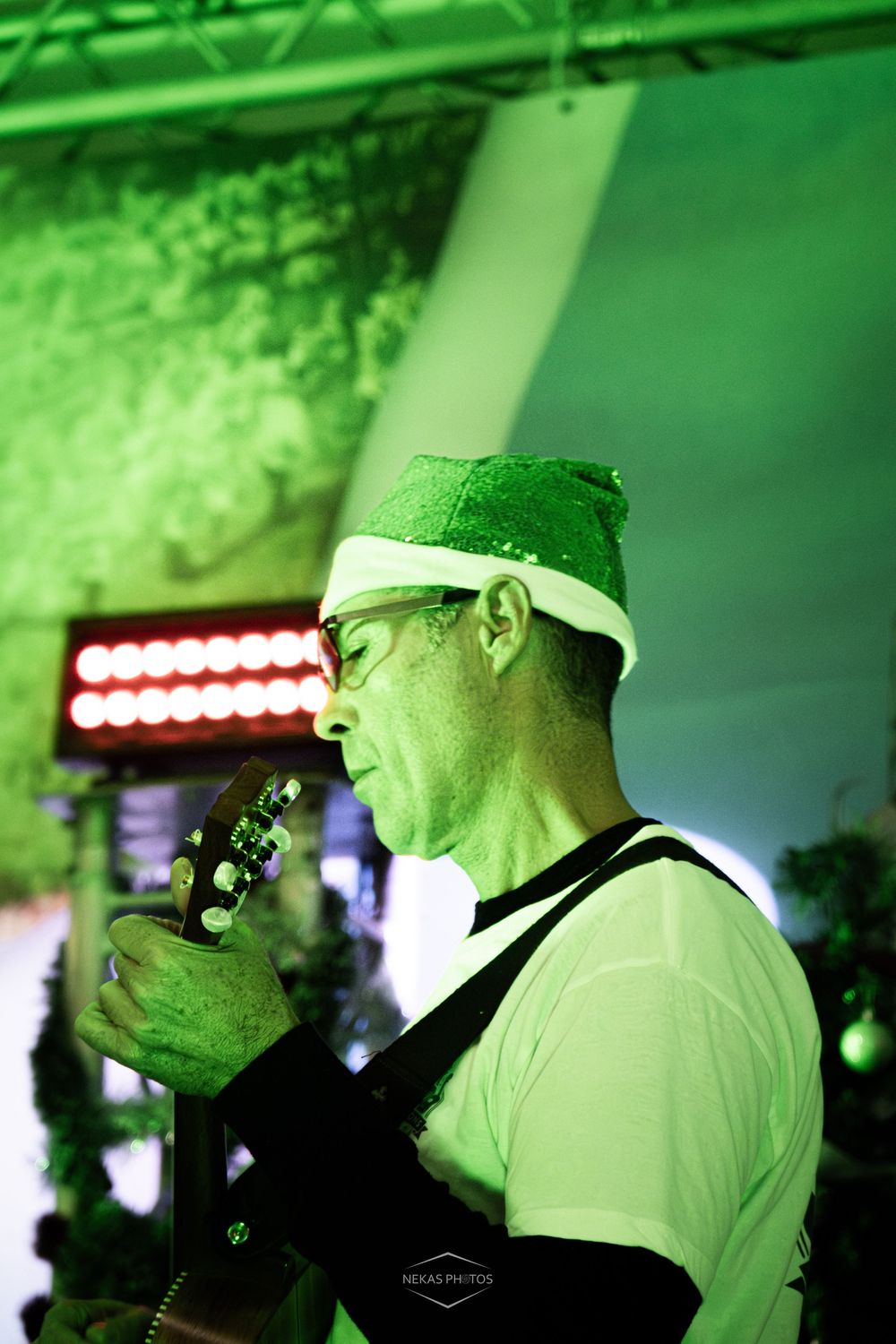 Musician in green sequin Santa hat plays guitar under vibrant green stage lighting at festive event.