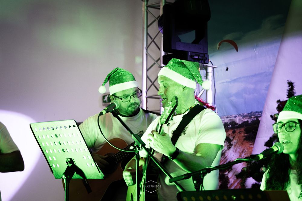 Musicians wearing Santa hats perform on stage under green lights at a festive Christmas concert.