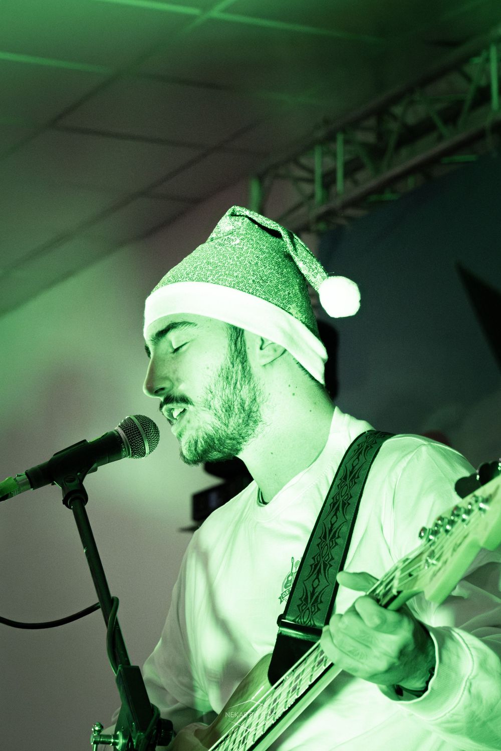 Bearded male guitarist wearing Santa hat performs on stage under green lighting at a Christmas concert.