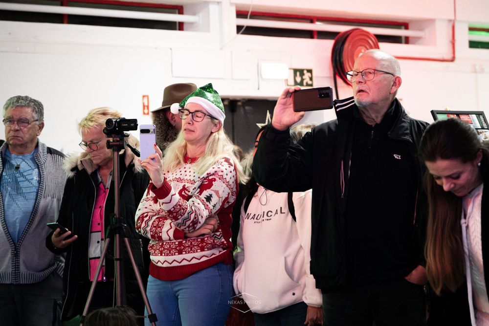 Group of people at a Christmas event, woman in festive sweater and elf hat stands beside man raising his hand.