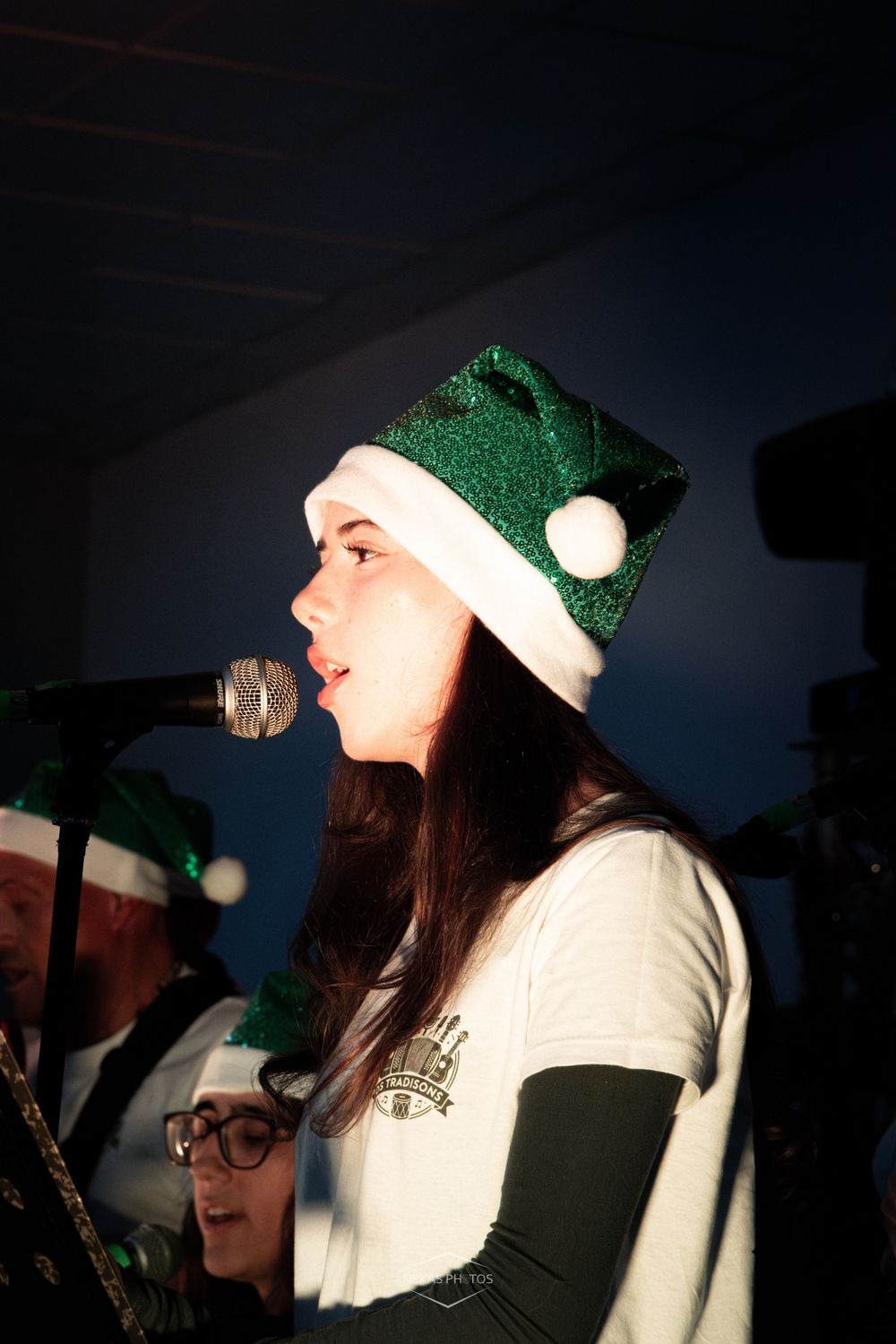 Young woman singing into microphone wearing festive green and white glittery Santa hat at holiday music performance.