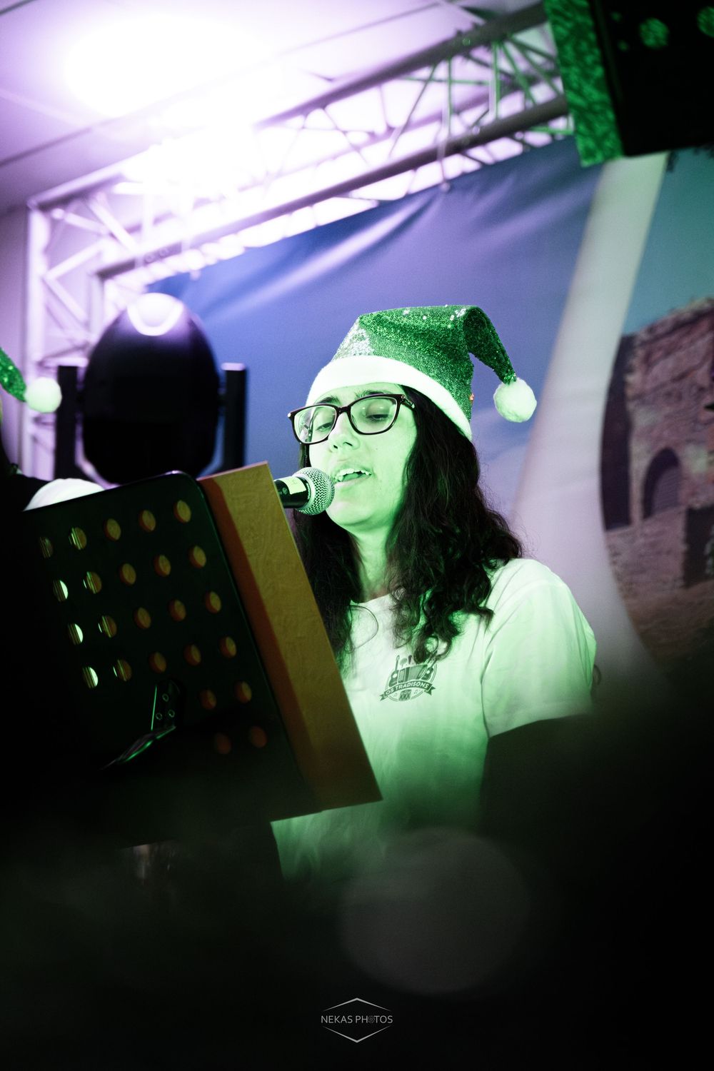 Woman wearing green Christmas elf hat and glasses singing into microphone at holiday concert event.