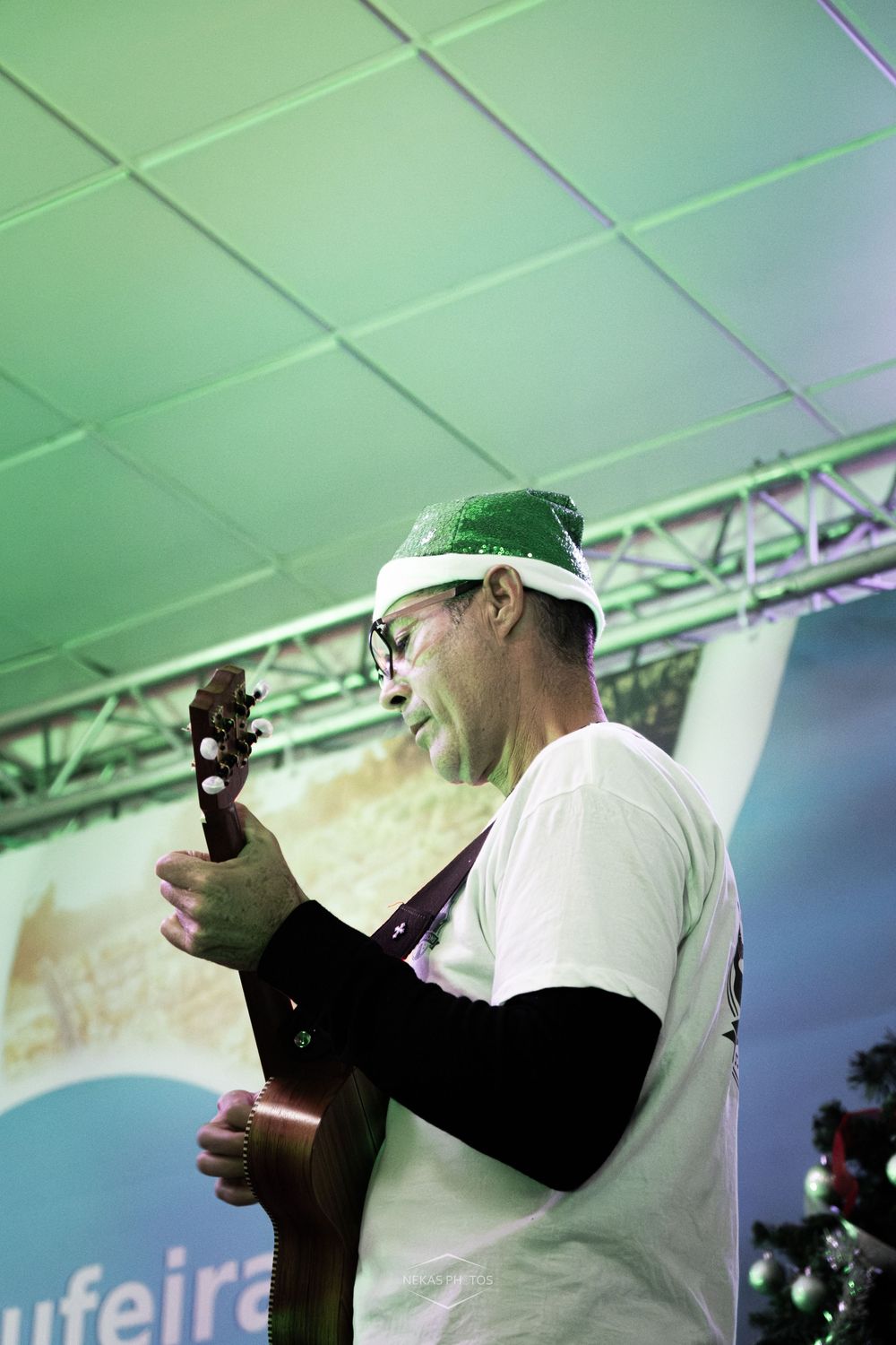 Man in festive green hat playing ukulele on stage at outdoor Christmas event.