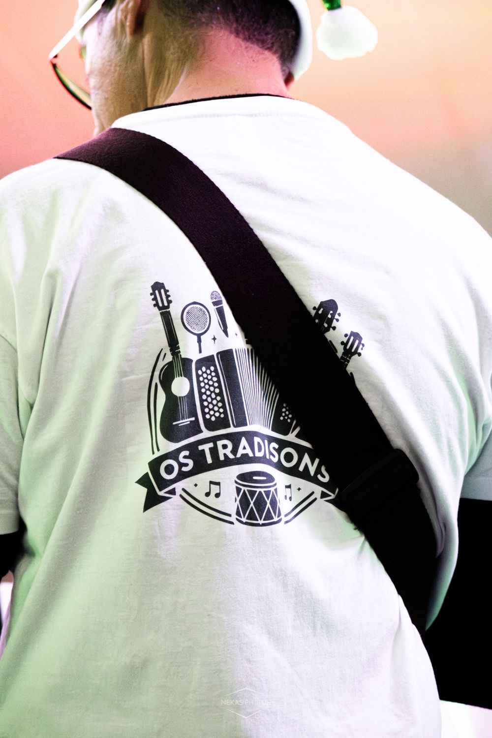 Man wearing white Os Tradisons band t-shirt with musical instruments logo, guitar strap across back.