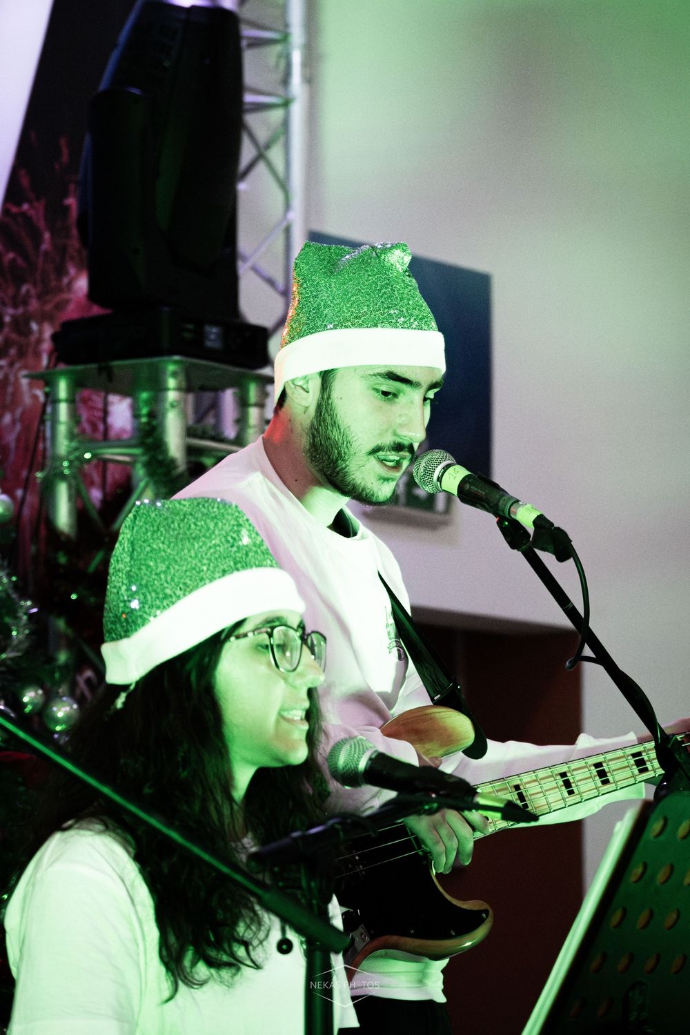 Two musicians in green Santa hats perform on stage, one playing guitar and singing into a microphone at a Christmas event.