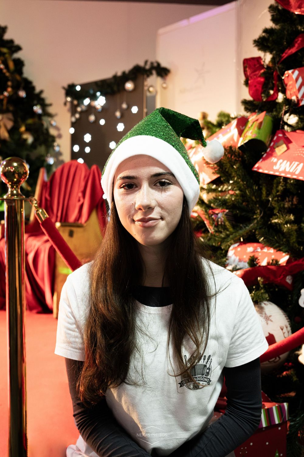 Young woman wearing a green and white elf hat smiling in front of a decorated Christmas tree with gifts.