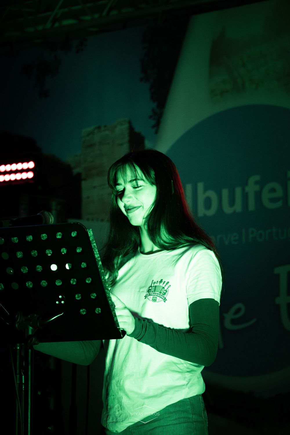 Young woman performing on stage under green lighting, reading from a music stand at an outdoor evening event in Portugal.