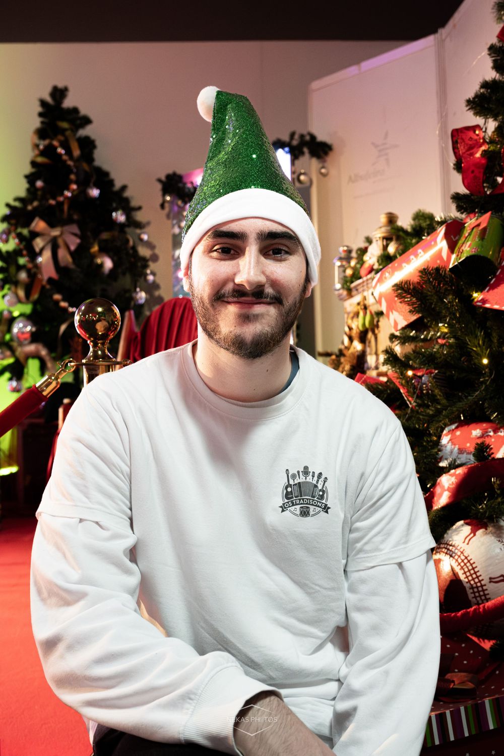 Man in green elf hat and white sweatshirt smiling in front of decorated Christmas trees.