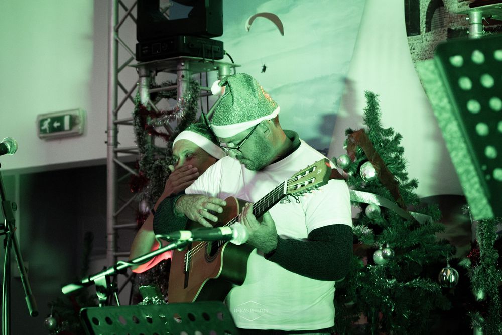 Musician in festive costume playing acoustic guitar on stage with green lighting and Christmas tree backdrop.