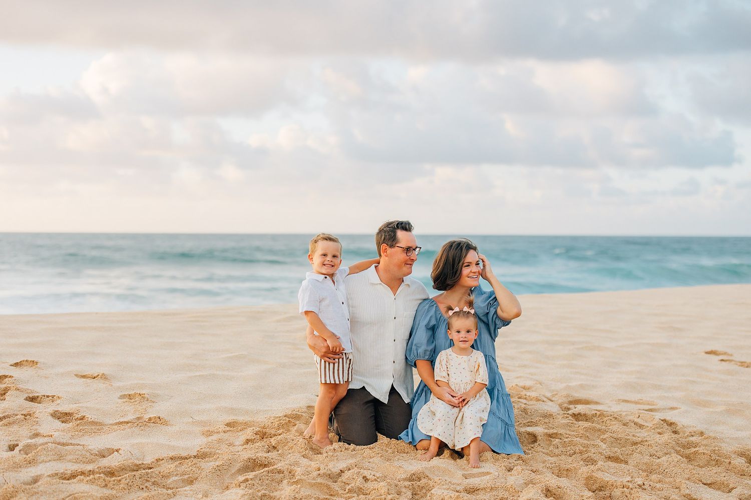 Family of four poses on sandy beach with ocean waves and pastel sky backdrop during golden hour.
