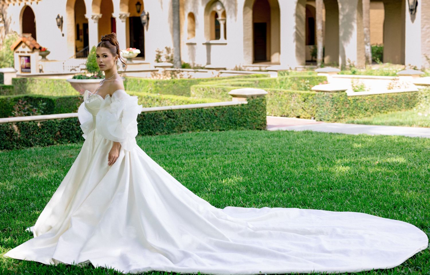 Bride in elegant off-shoulder white ball gown with long train poses gracefully in lush garden setting.