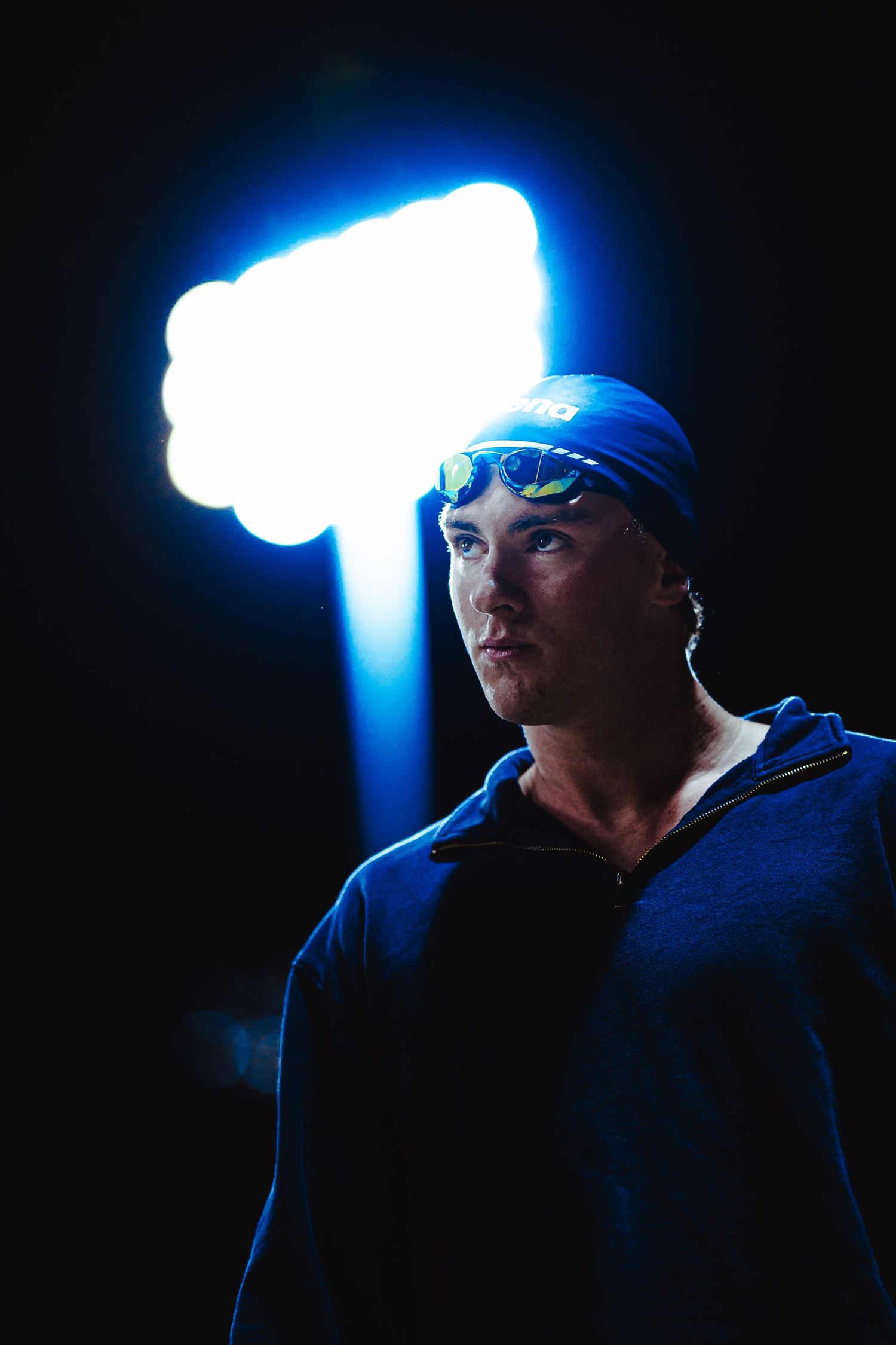 Male swimmer in white cap and black swimsuit stands poolside at an outdoor competitive swimming event.