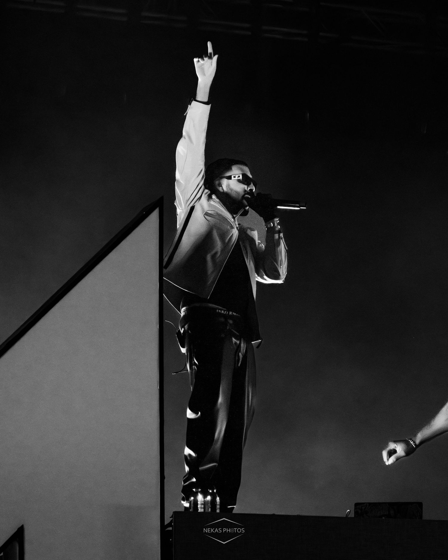 Black and white photo of a performer on stage holding a microphone with one arm raised high at a live concert.