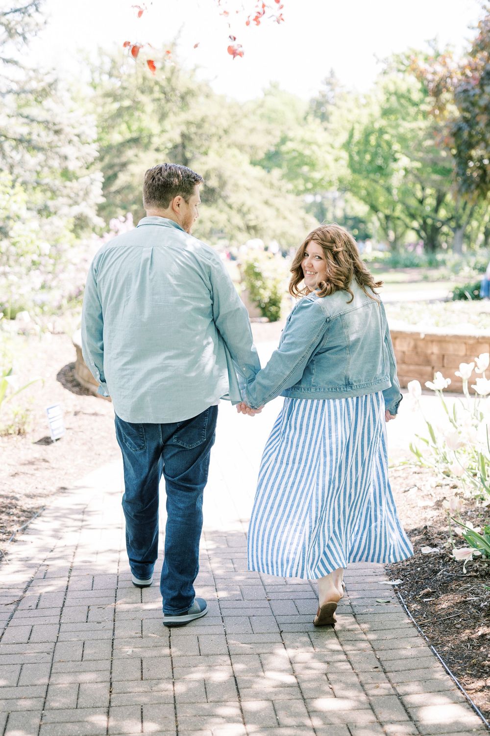 Couple walking hand in hand on a brick path, woman in striped skirt glancing back smiling.